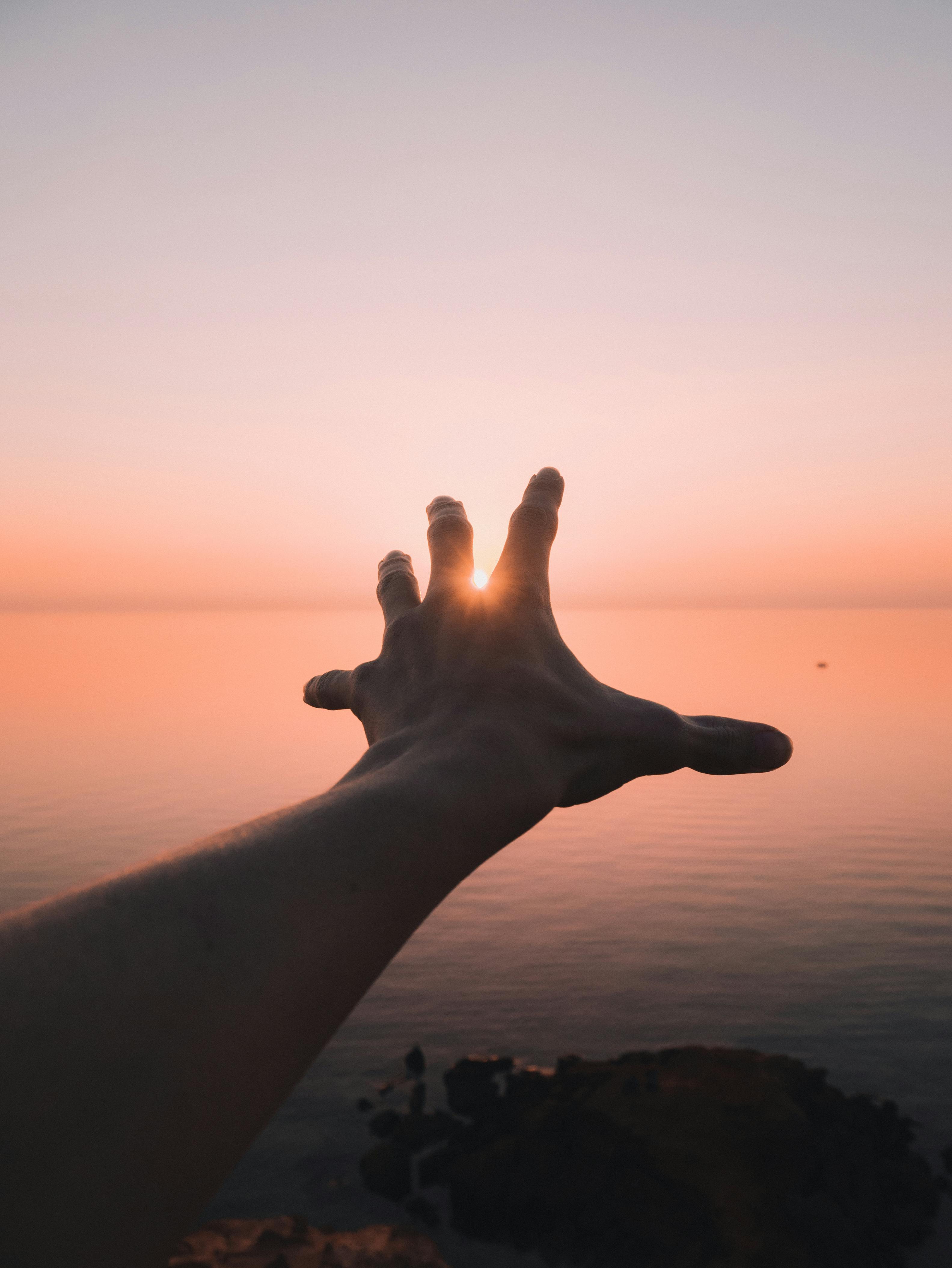 A silhouette of a hand reaching towards the setting sun over a tranquil ocean at Black Rock, Australia.