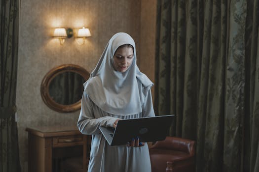 Woman wearing a hijab using a laptop indoors, showcasing work and technology.