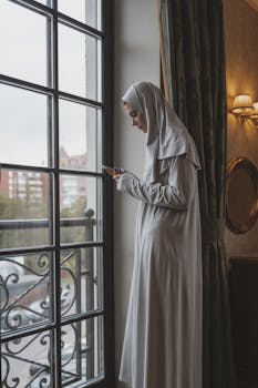 A Muslim woman in hijab looking at her phone by a window indoors.
