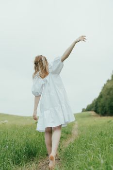 一名妇女赤脚走在郁郁葱葱的绿草地上，她身着白色连衣裙，步履平静