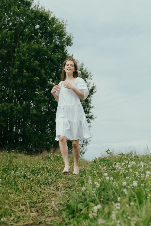 Free A woman in a white dress walks barefoot in a grassy meadow during summer. Stock Photo
