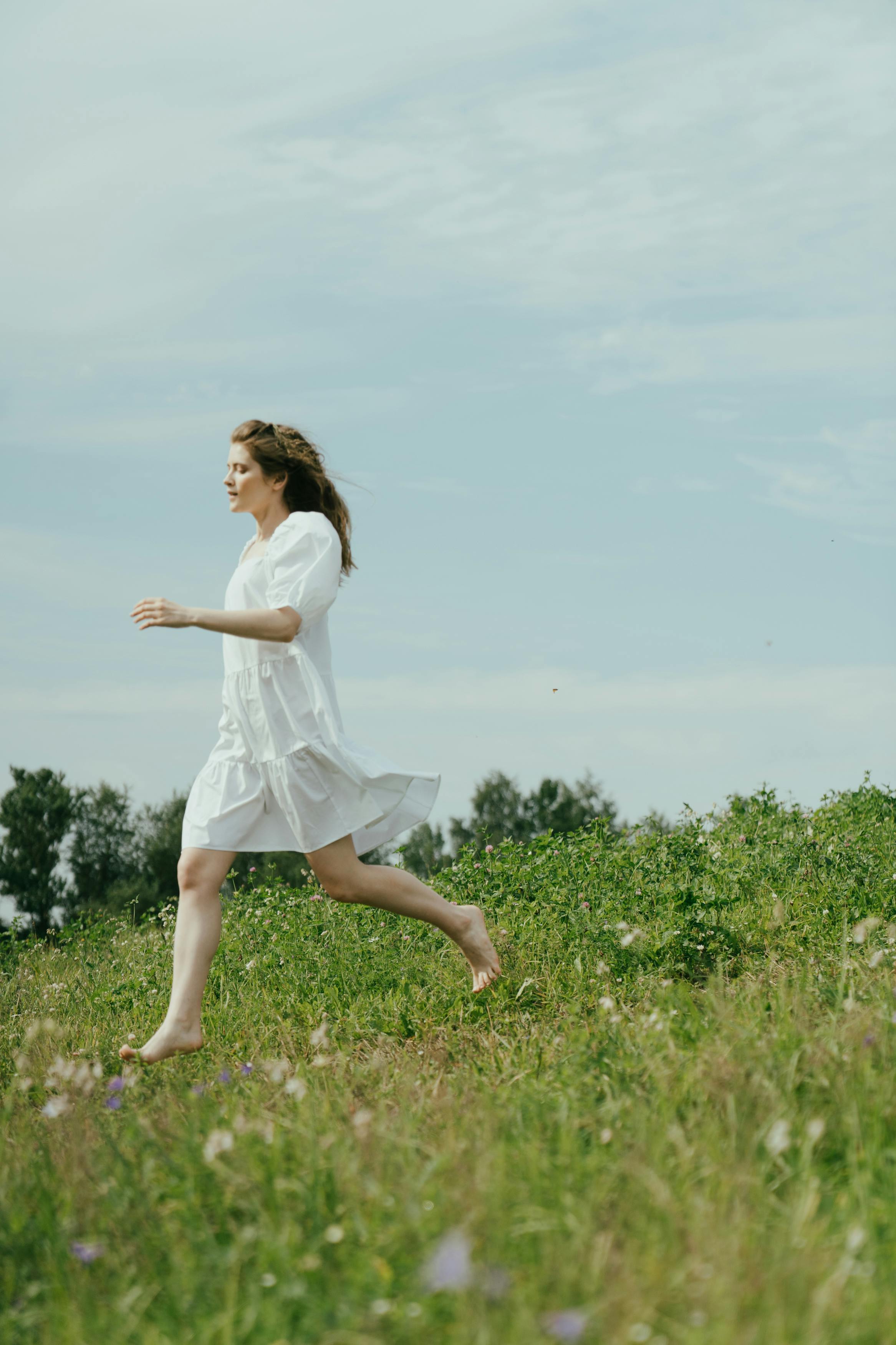 一个穿着白色连衣裙的女人在晴朗的天空下快乐地赤脚穿过绿色的草地