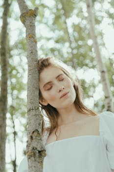 一位身着白色连衣裙、闭着眼睛的宁静女子，正靠在户外的树干上