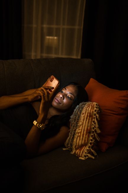 Woman relaxing on a sofa at night, using a smartphone, surrounded by cozy pillows.