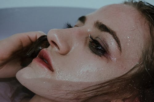 Young female with wet face touching hair in clean transparent water of bathtub