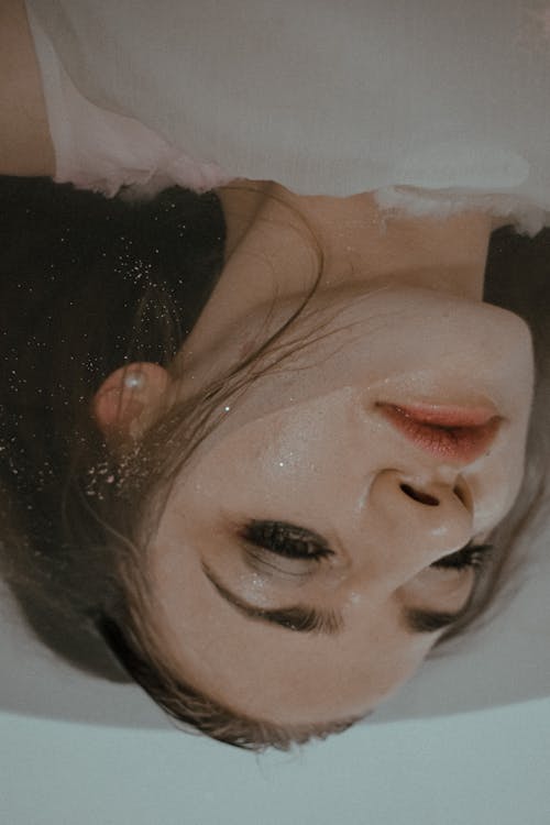 Unhappy female with eyeliner and drops on face resting in water in bathtub