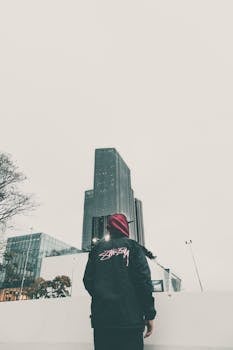 Individual in streetwear looks at city skyscraper, showcasing urban fashion against a modern cityscape.