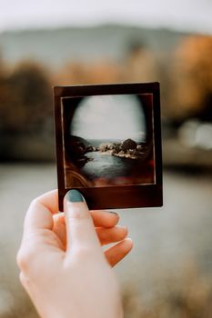 A hand with blue nail polish holding a Polaroid picture of a river landscape, in an autumn setting.