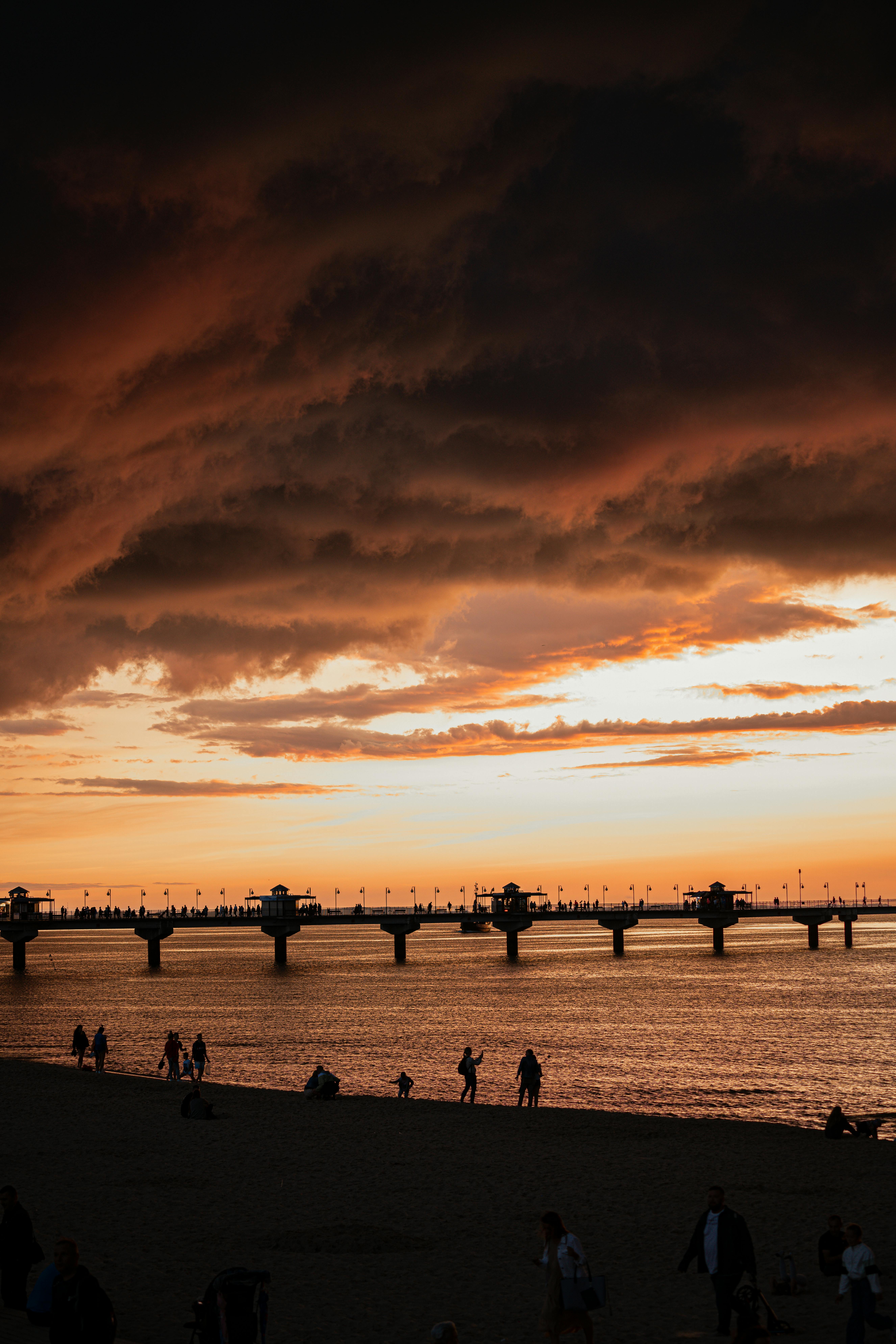 A stunning sunset over a pier in Miedzyzdroje, Poland, with silhouettes of people and dark clouds.
