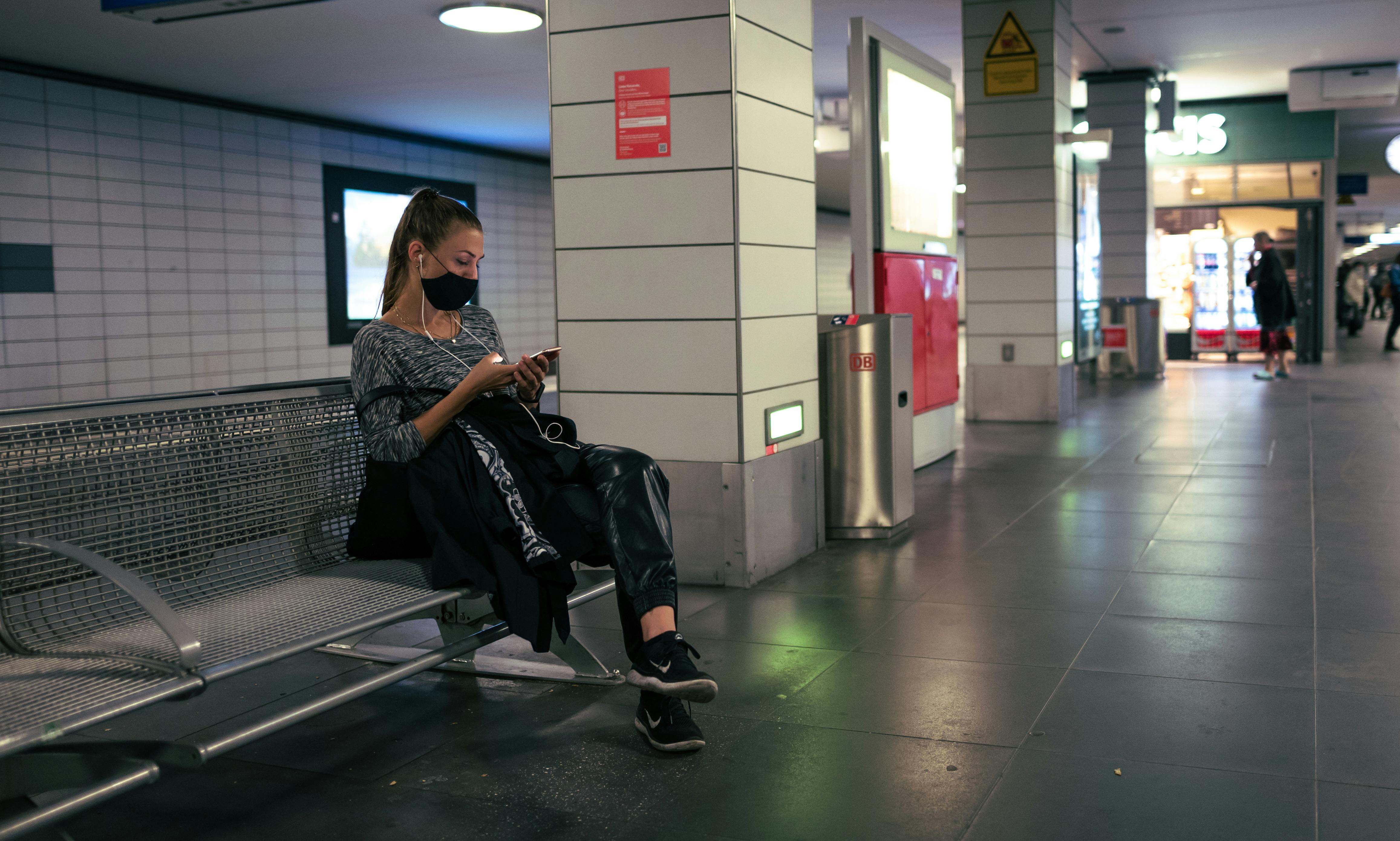 Woman sits on a bench in a subway station, wearing a face mask and using her phone.