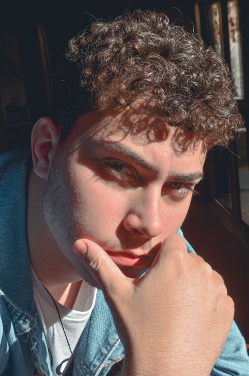 Free Close-up portrait of a young man with curly hair pondering. Warm indoor setting highlights his thoughtful expression. Stock Photo