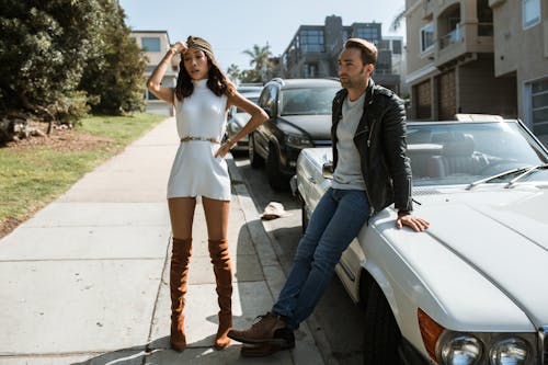 Woman in White Dress and Man in Black Leather Jacket on Sidewalk Near Cars