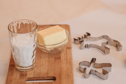 A glass of milk and butter bowl placed on a wooden board with cookie cutters, ready for baking.