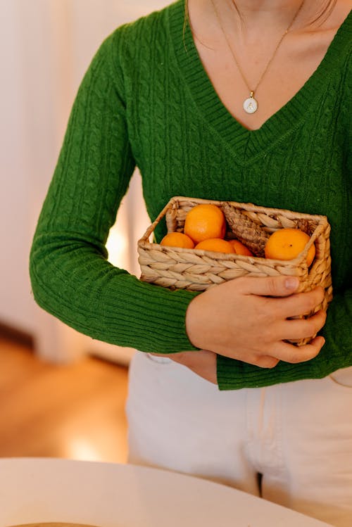 Person in Knitted Long Sleeves Carrying a Woven Basket with Orange Fruits 