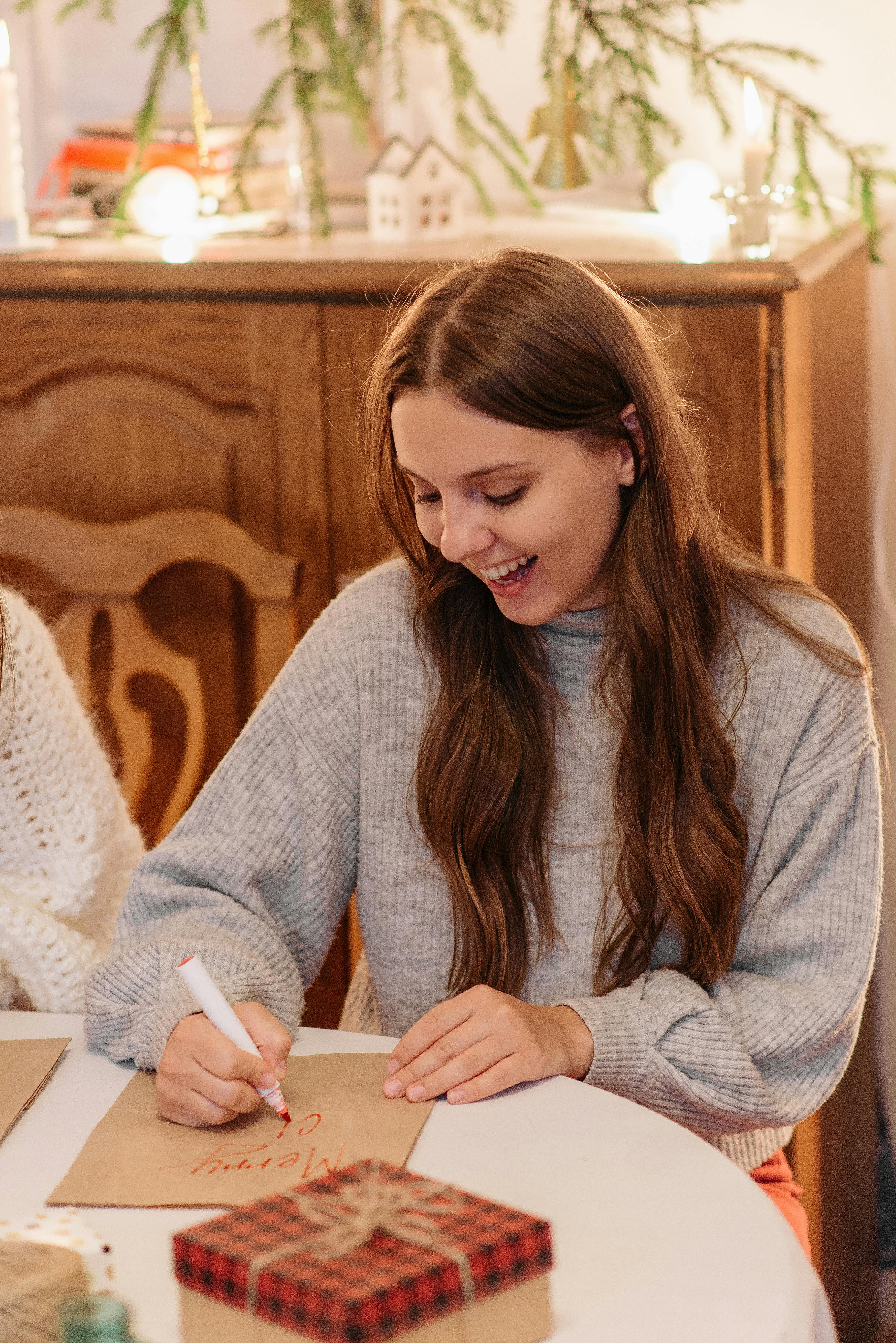 一位穿着灰色毛衣的快乐女人在摆满礼物和装饰品的节日餐桌旁写着贺卡