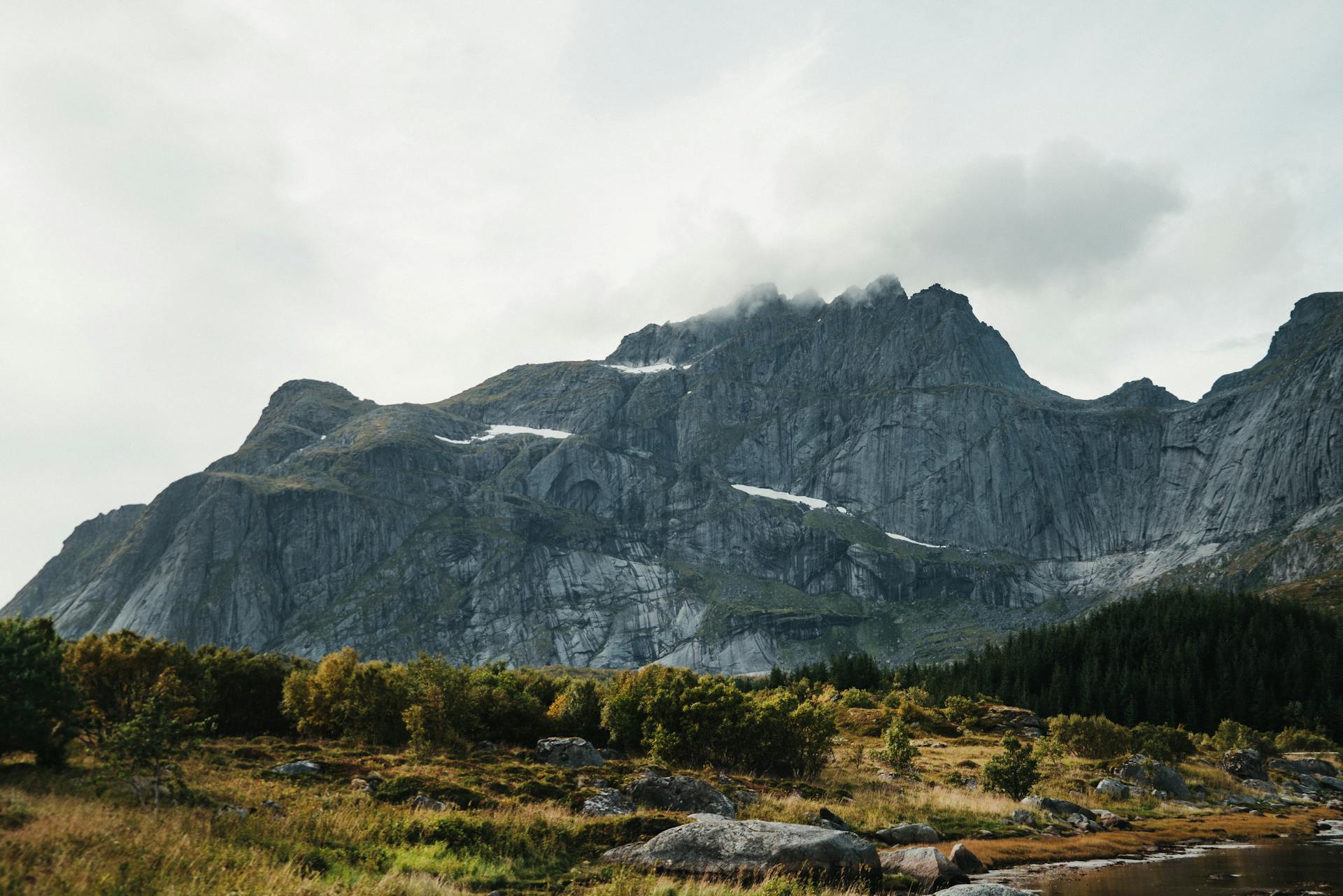 Majestic huge range of gray rocky mountains with snow high in clouds and green terrain below