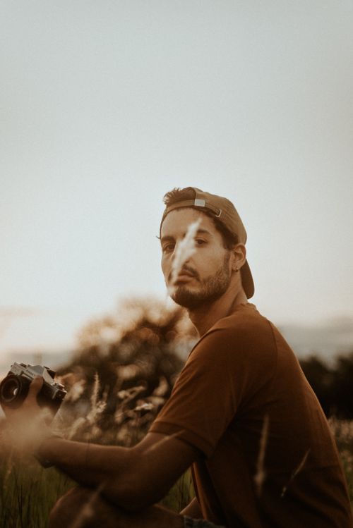 Free A young man in a cap with a camera, enjoying a serene outdoor scene during golden hour. Stock Photo