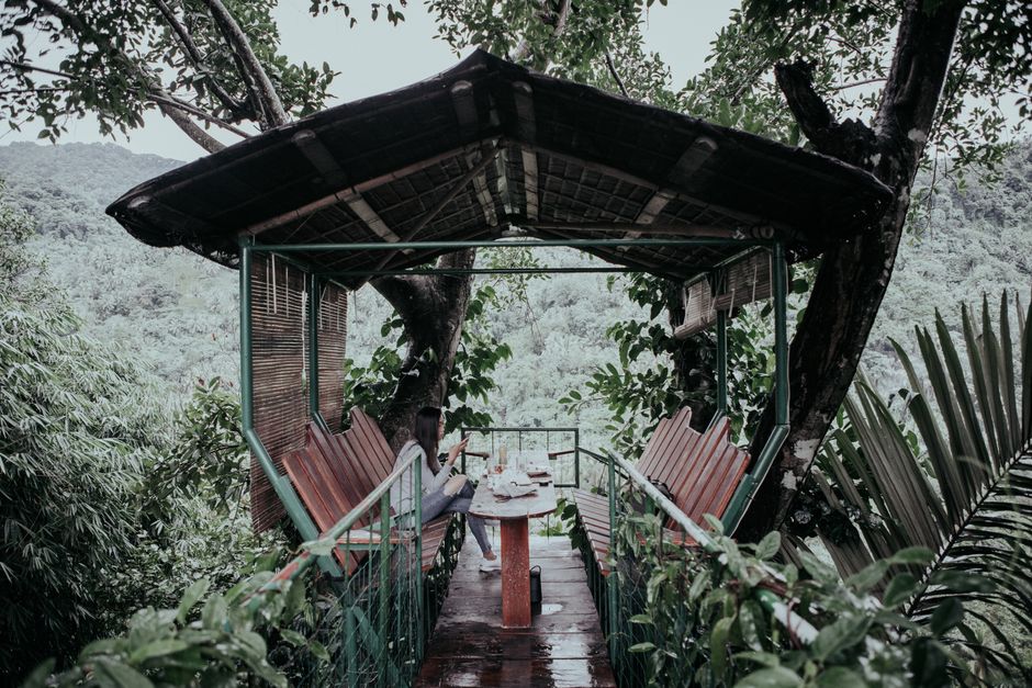 Side view of unrecognizable female traveler sitting with crossed legs on bench in aged arbor while looking away near green mounts