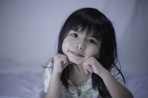 Free Smiling ethnic child with tilted head sitting leaned on hands on bed while looking at camera in house Stock Photo