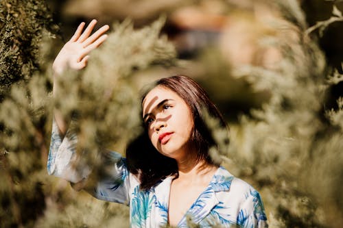 Young dreamy Asian female in stylish clothes with makeup standing with raised arm among greenery shrubs while looking up