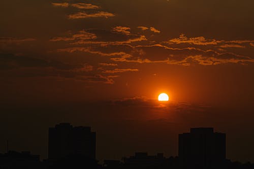 Majestic scenery of clouds floating on sundown sky above roofs of buildings with distant sun setting on horizon in evening time