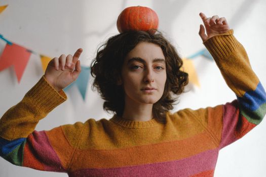 Woman with curly hair balancing a pumpkin on her head. Fall-themed indoor scene with colorful flags.