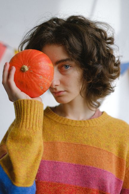 A young woman with curly hair holds a pumpkin, displaying fall fashion in a colorful sweater.