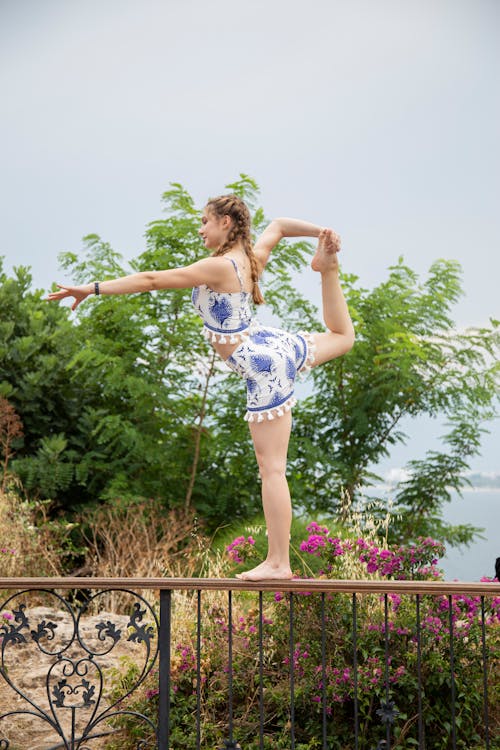 Side view of flexible barefoot teen in trendy wear raising leg on metal fence under white sky while looking forward