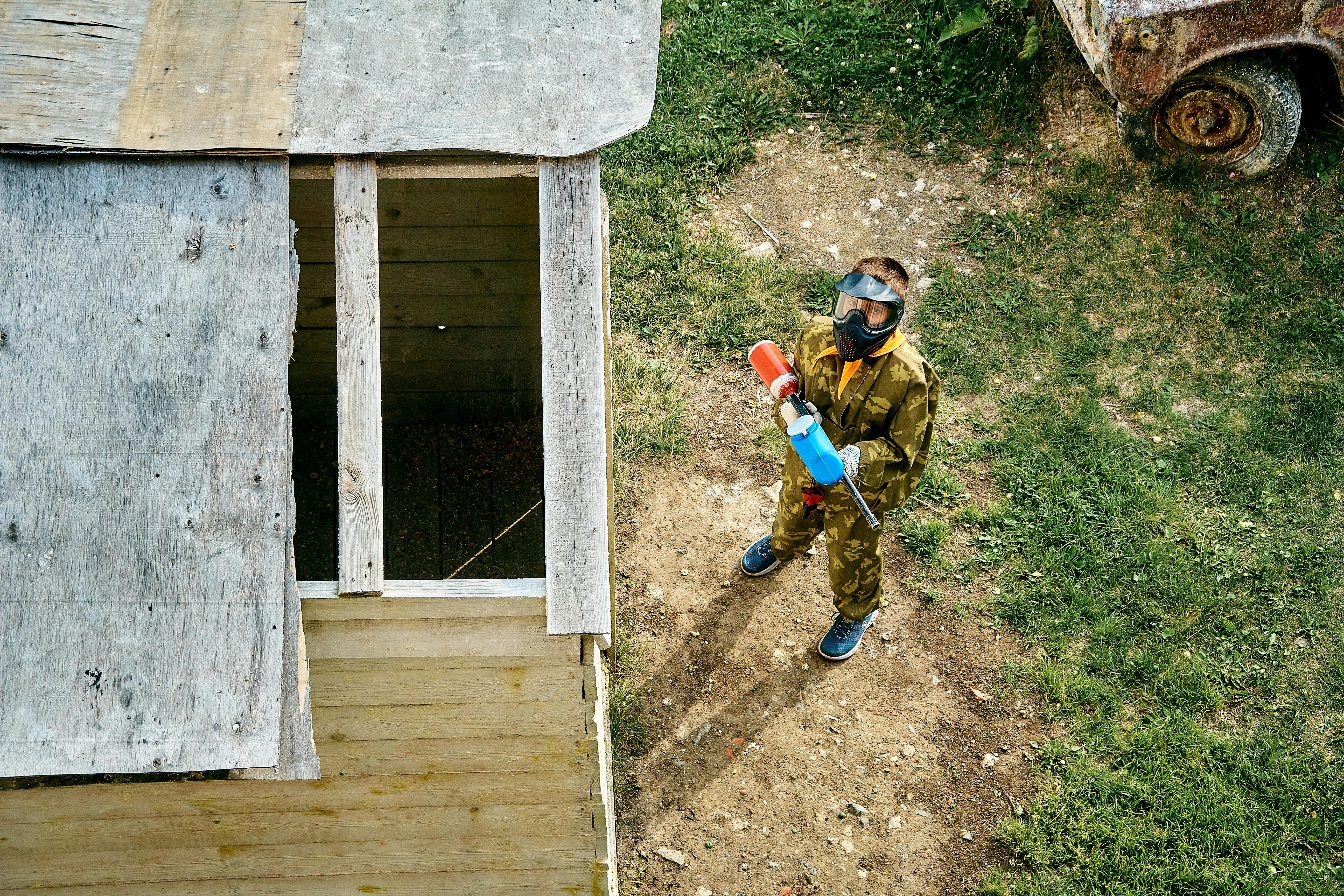 Aerial view of a young person in full gear playing paintball outdoors next to a wooden structure. Prescription Goggle Inserts - Aerial view of a young person in full gear playing paintball outdoors next to a wooden structure.