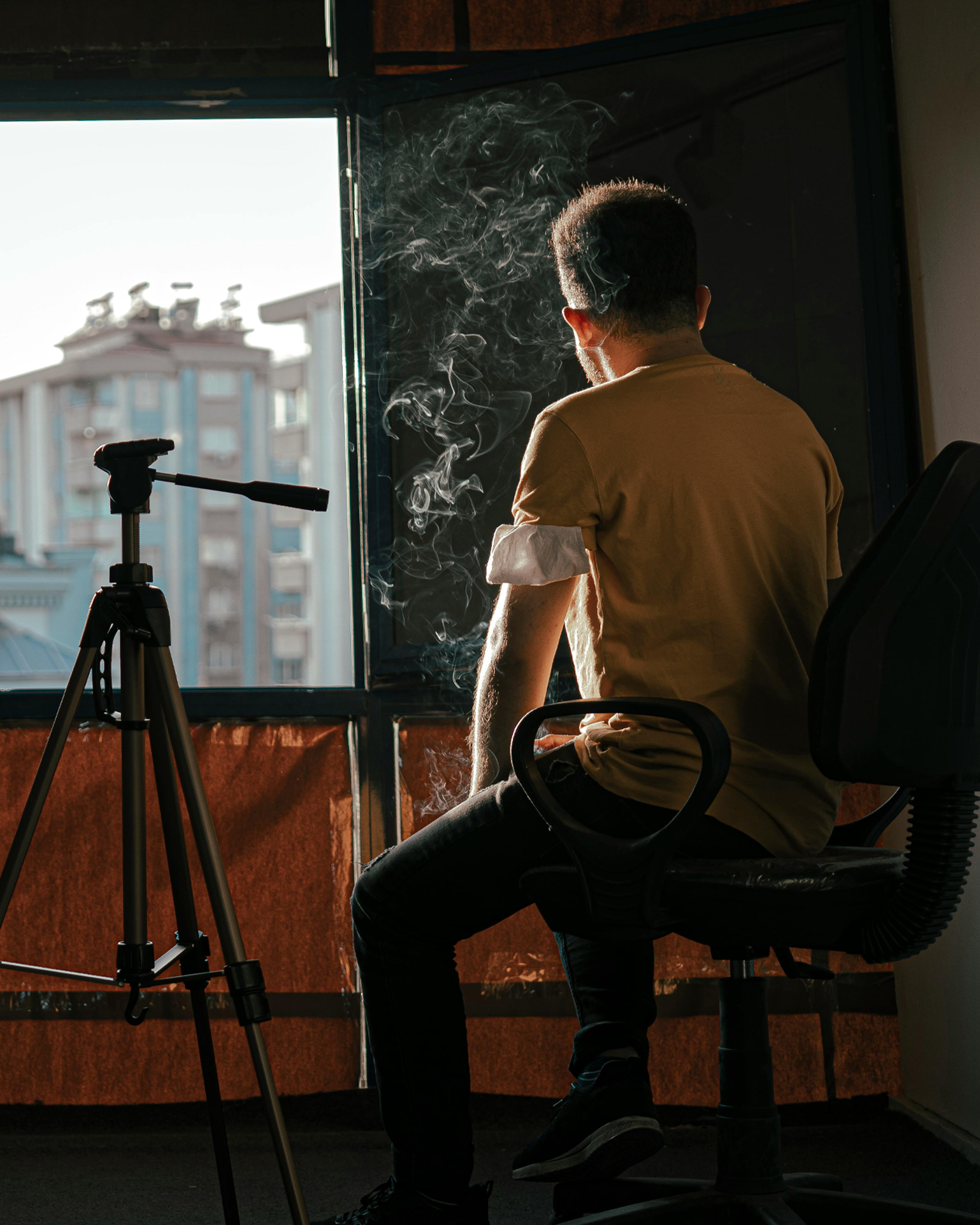 A man sits near a window with smoke and a tripod, capturing a moment of contemplation.
