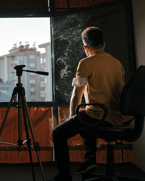 Free A man sits near a window with smoke and a tripod, capturing a moment of contemplation. Stock Photo