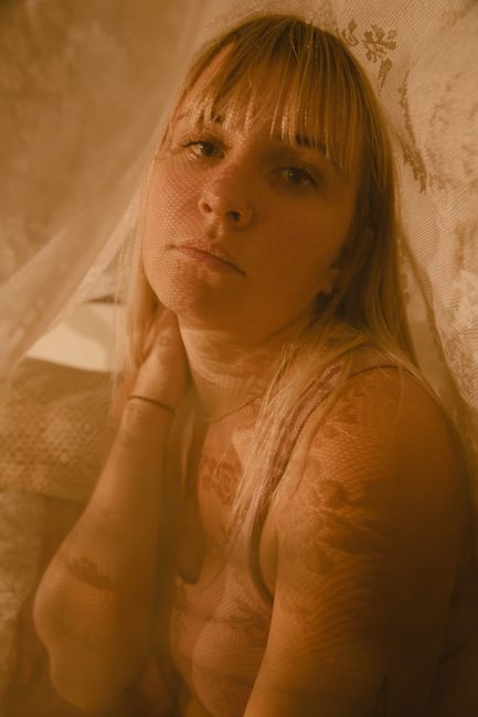 A thoughtful young woman with bangs poses under lace fabric, creating a soft and intimate mood.
