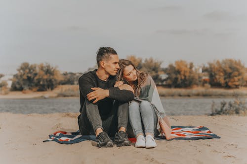 Full body of loving couple hugging and looking away while sitting on blanket with bent legs on sandy beach near calm lake in nature with blurred background