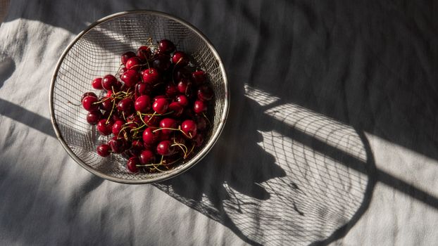 From above of heap of colorful tasty cherries with peduncles in metal strainer on crumpled textile with shadow
