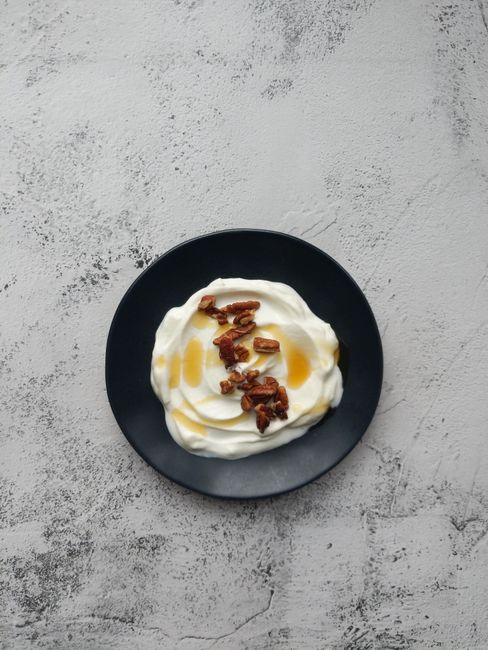 A glass jar of thick, strained vanilla Greek yogurt with a honey drizzle on top and a spoon resting beside it.