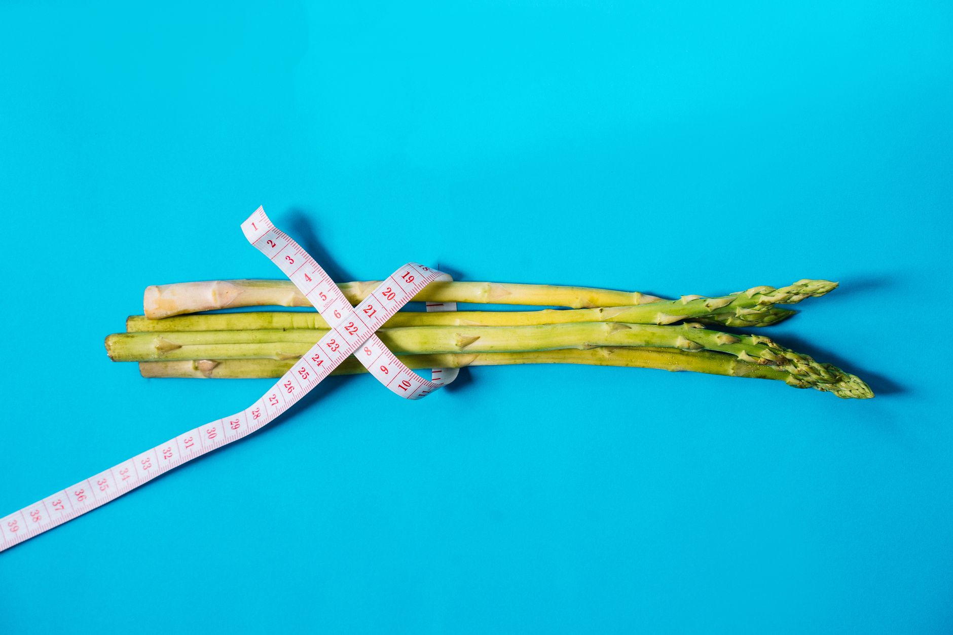 三種減重法大比拼:優缺點與適合族群 Fresh asparagus wrapped with measuring tape on a vibrant blue background.