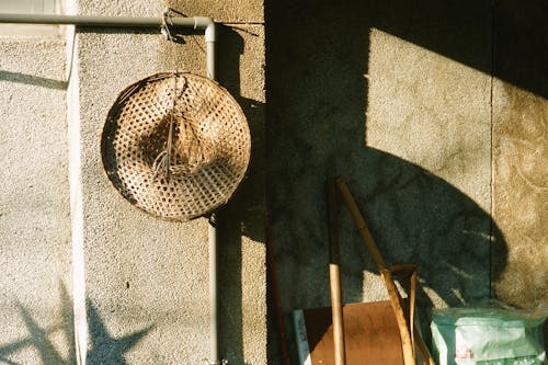Straw hat hanging on pipe near concrete wall with shadow near junk yard in sunny weather in street in daylight