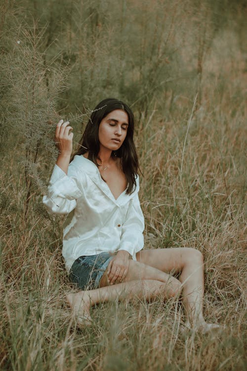 Side view of calm barefooted young ethnic lady in casual outfit resting in hay with closed eyes in countryside