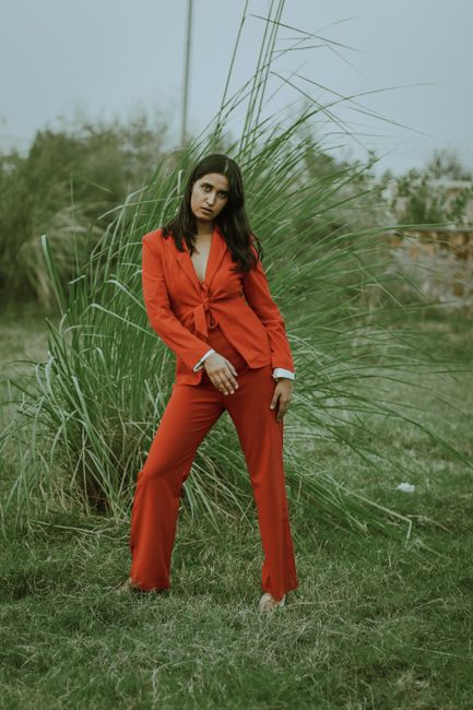 Full body of young confident ethnic female in stylish red suit standing on grassy lawn near green bush and looking at camera