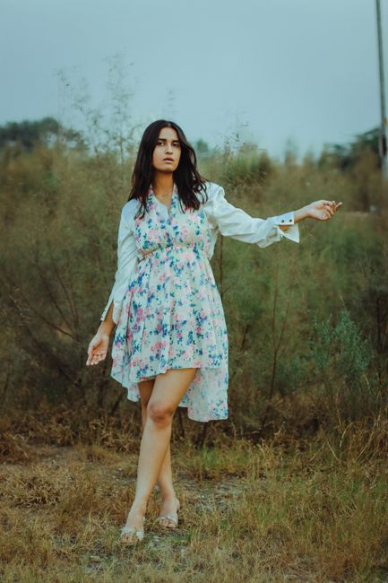 Full length of alluring young ethnic brunette in light dress relaxing on dry grassy meadow and looking at camera against cloudy sky