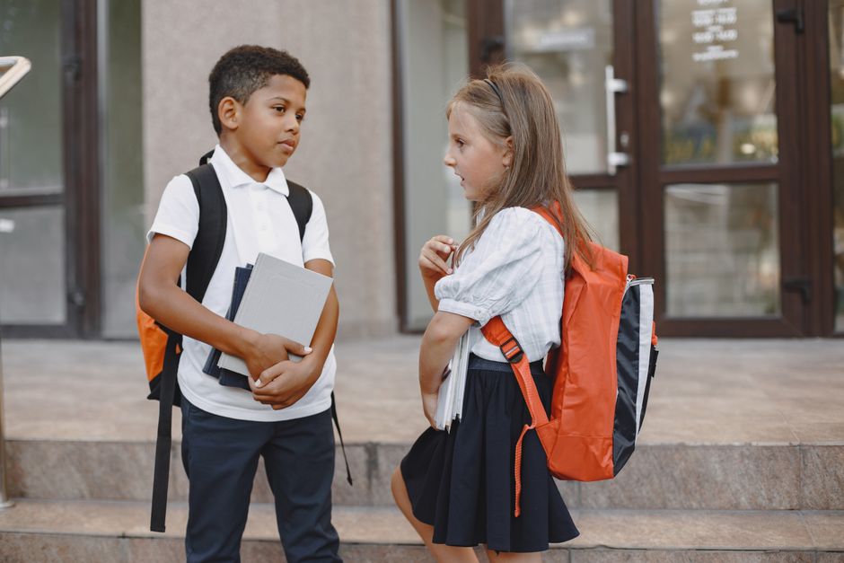 Two children in school uniforms holding books and talking outdoors.