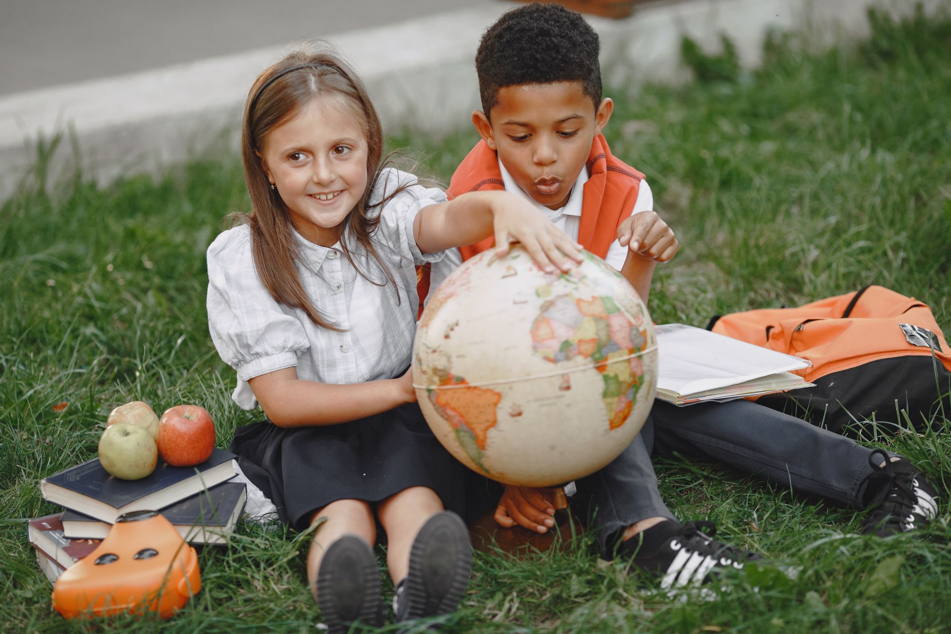 Two children in school uniforms exploring a globe, sitting on the grass and learning outdoors.