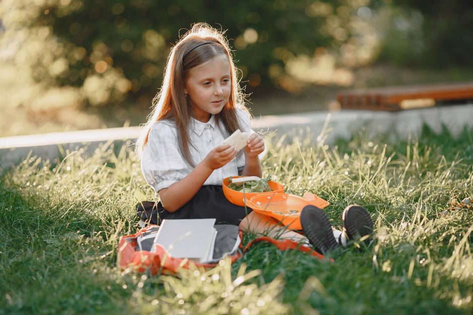 A young girl sitting on grass in a park, enjoying a sandwich during a sunny day picnic.