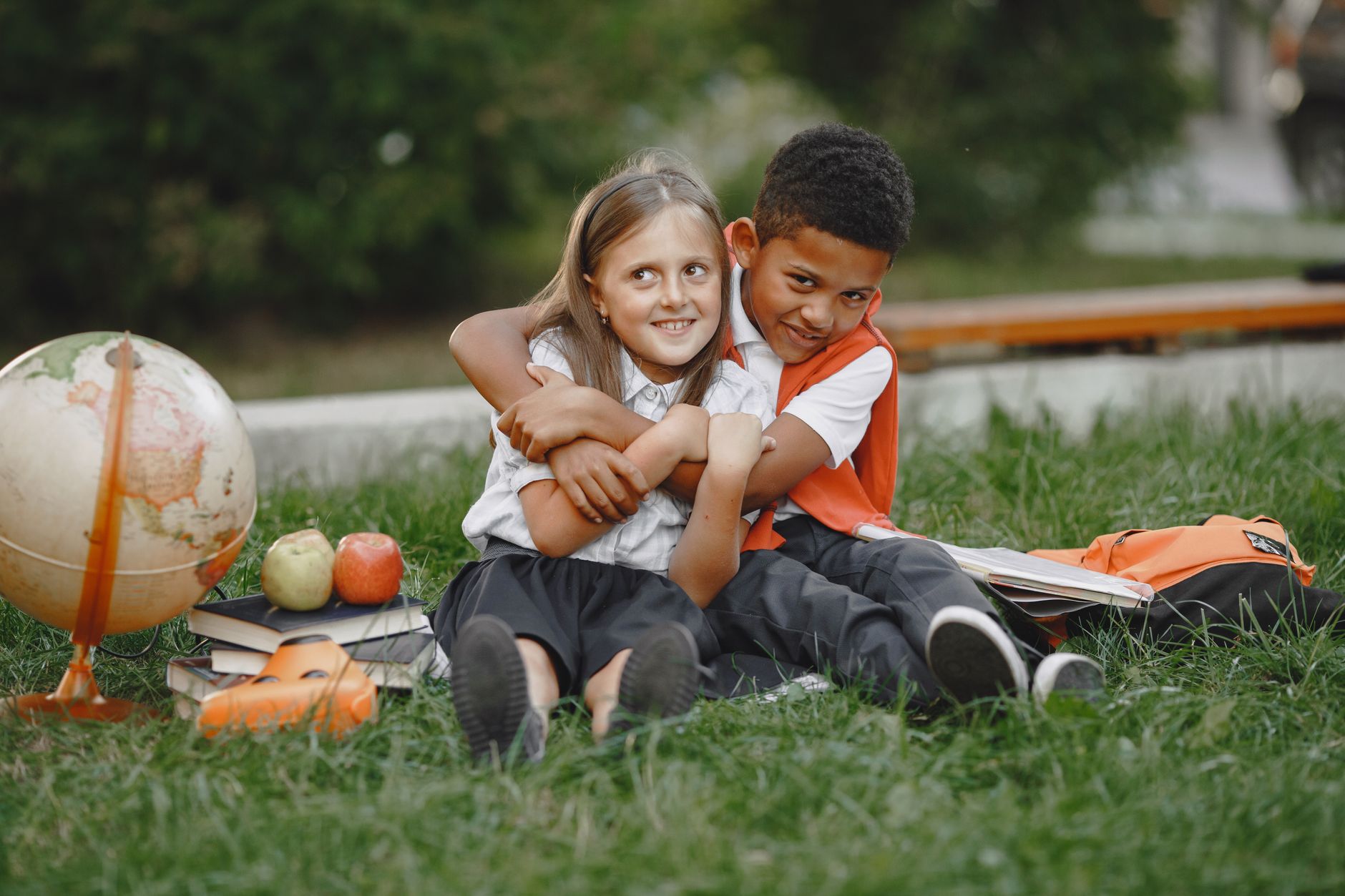 Two diverse kids in uniforms hugging outdoors with books and a globe, embodying education and friendship.