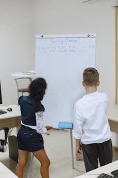 Two students work together on a whiteboard in a classroom setting.