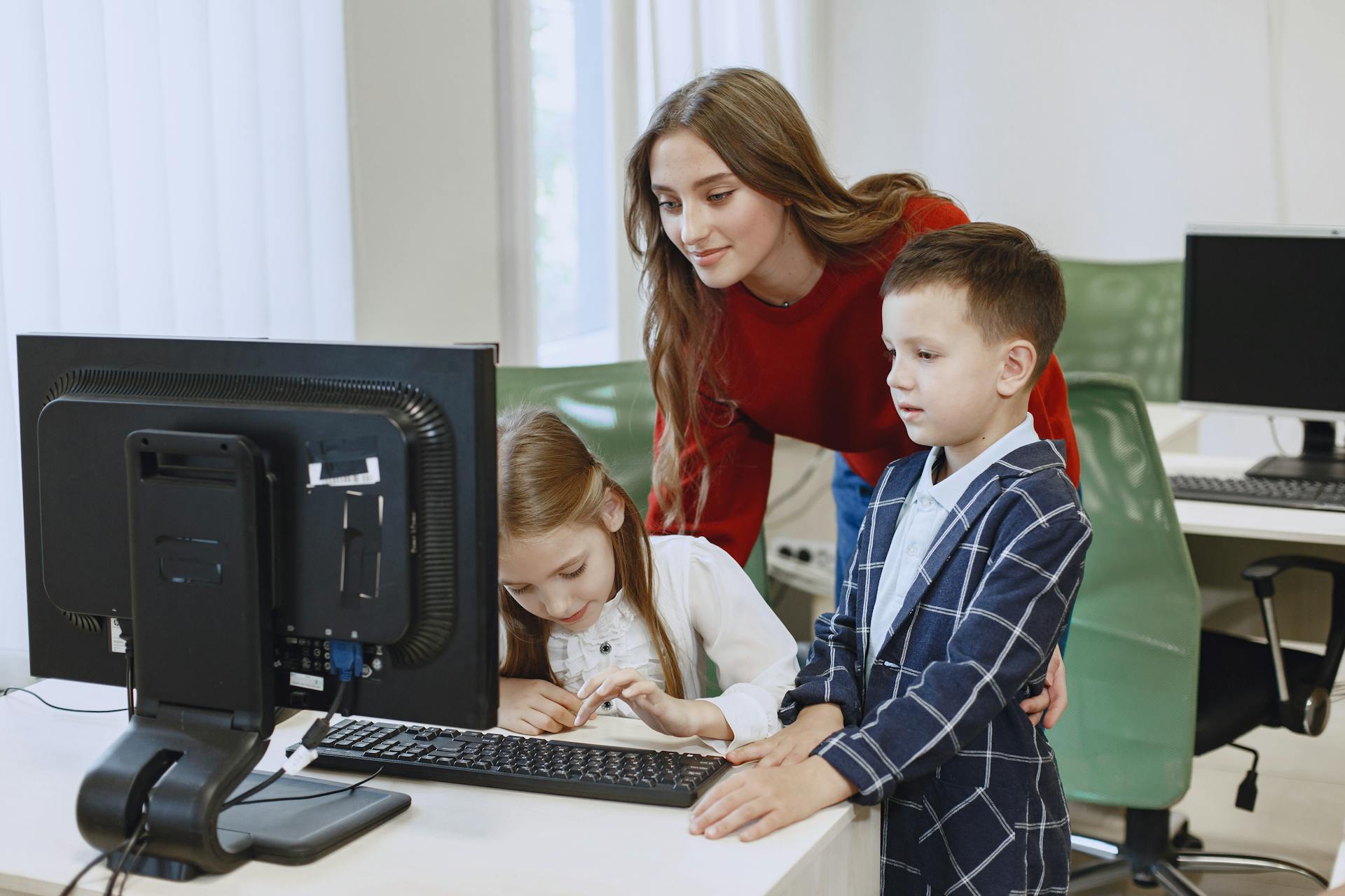 A teacher guides two children using a computer in a classroom setting.