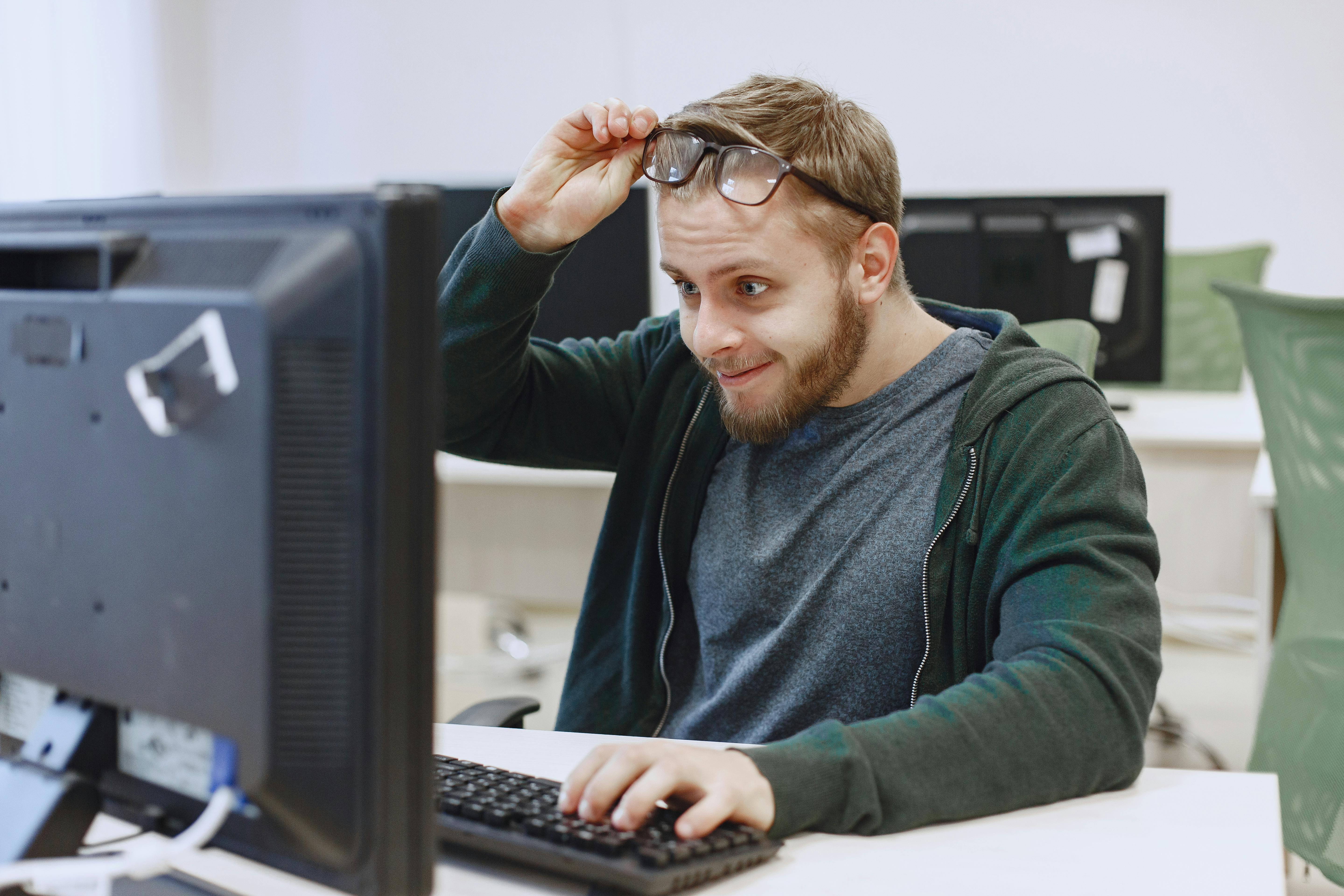 A surprised bearded man sits at a computer, lifting his eyeglasses in an office setting.