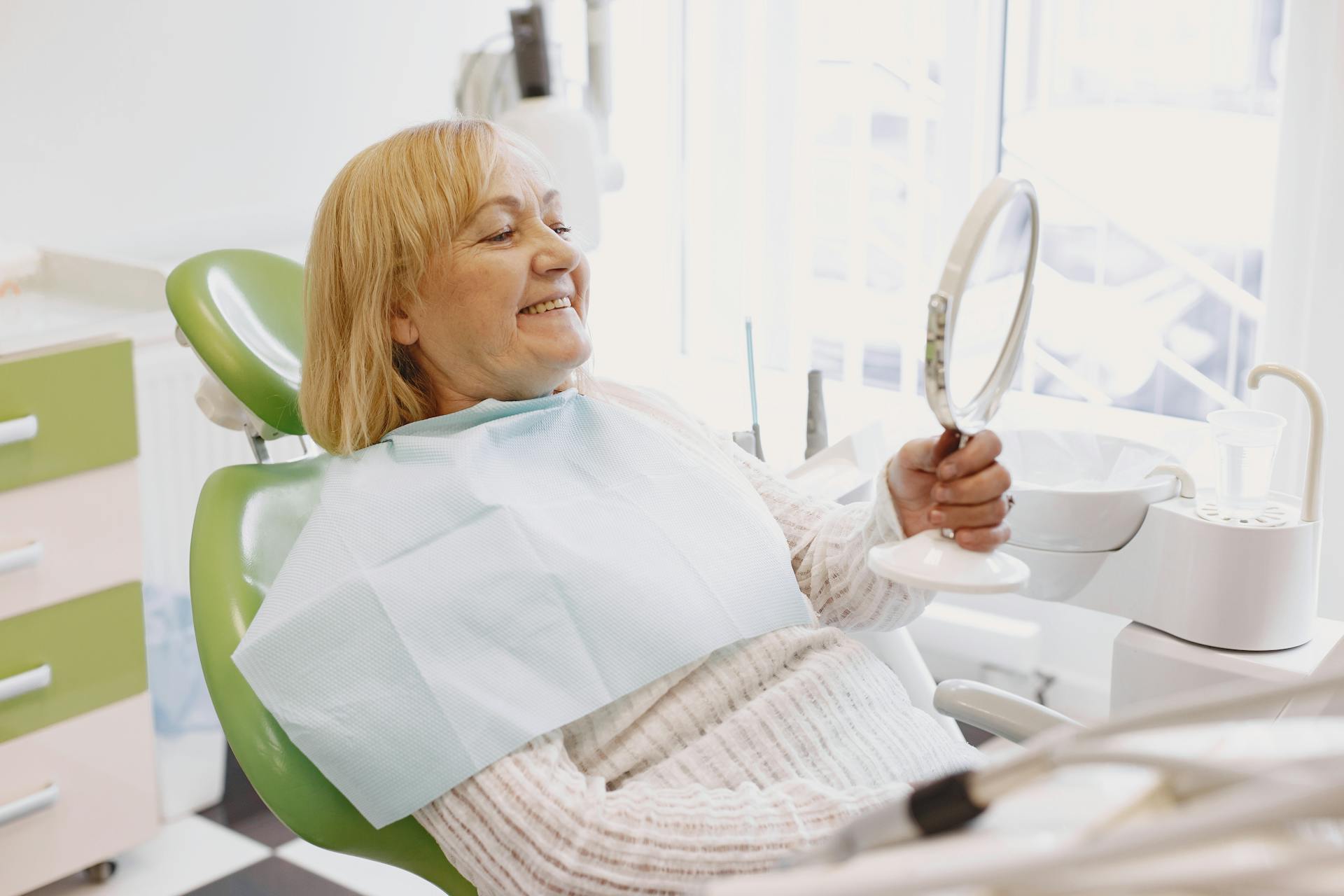 Elderly woman in dentist chair smiling while holding a mirror during a dental check-up.