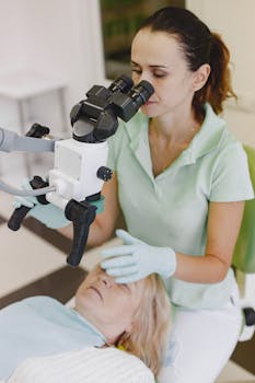 A female doctor in gloves examines a patient using a microscope in a clinical setting.