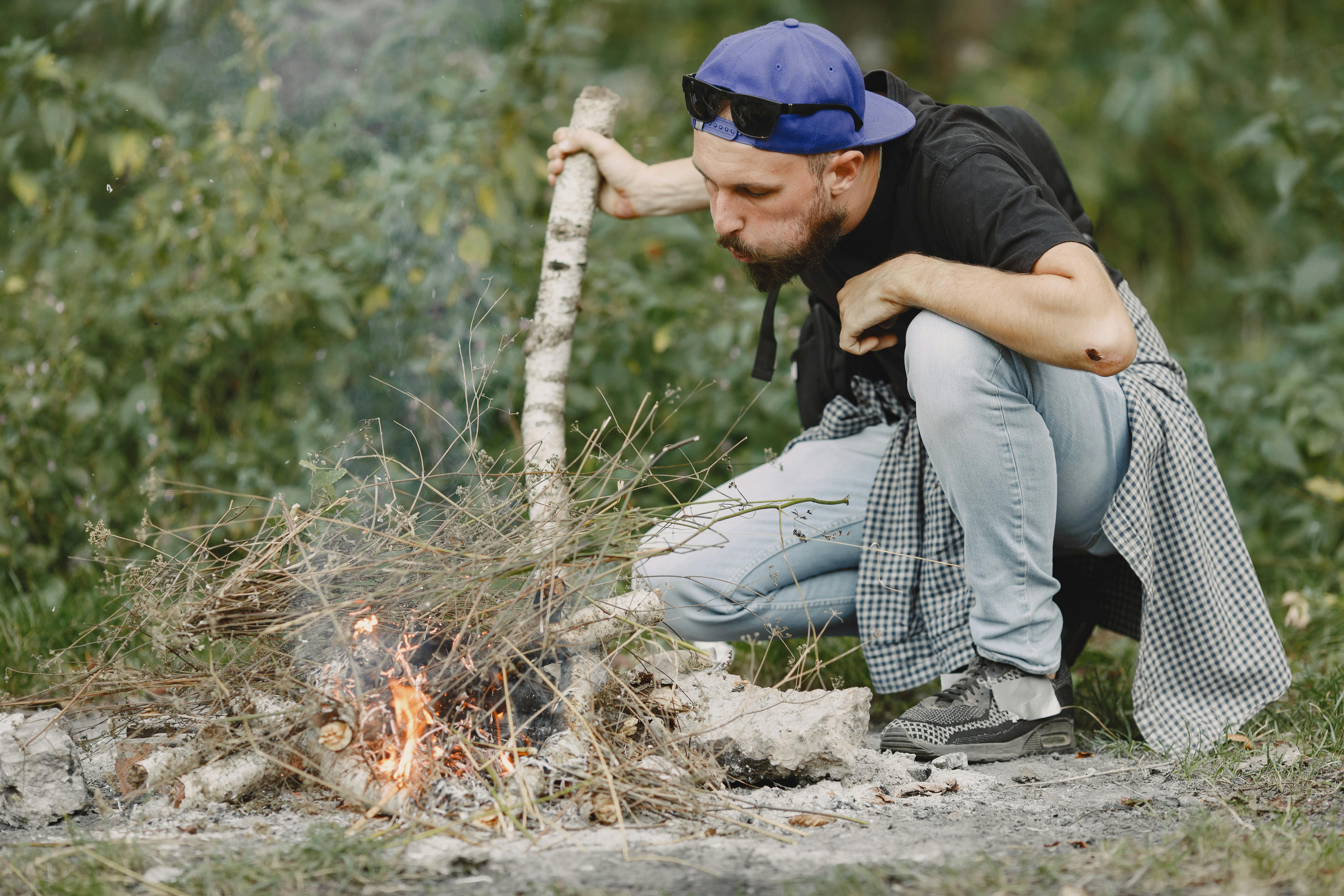 An adult man blowing on a campfire in the forest while wearing a blue cap and sunglasses.